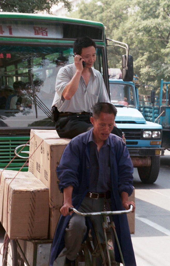 a man peddling a load of boxes with a man sitting on top